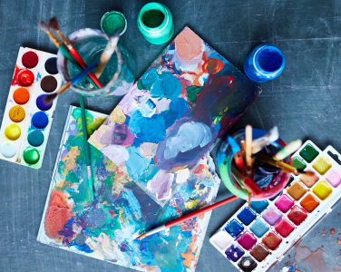 Colourful paper, paintbrushes in jars, and paint palettes. 