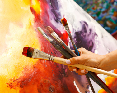 A hand holding paintbrushes in front of a painting with vibrant colours. 