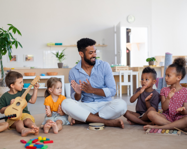 A teacher and children sitting on the floor, clapping and playing instruments.