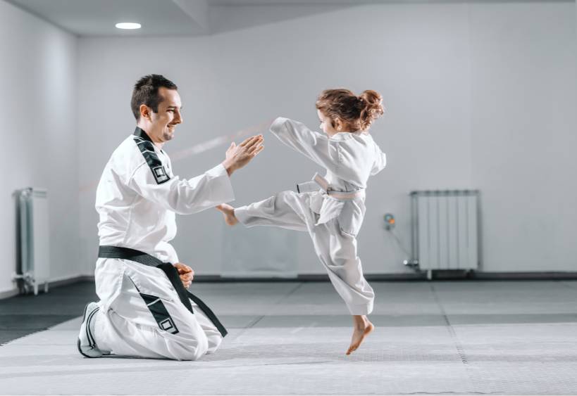 Smiling Caucasian taekwondo trainer in dobok kneeling and holding hand up while little girl kicking him.