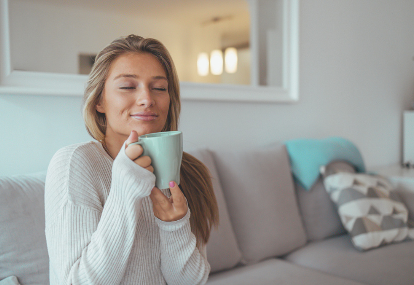 A calm, content woman sits on a gray sofa, holding a mint mug and enjoying a warm drink in a bright, cozy living room