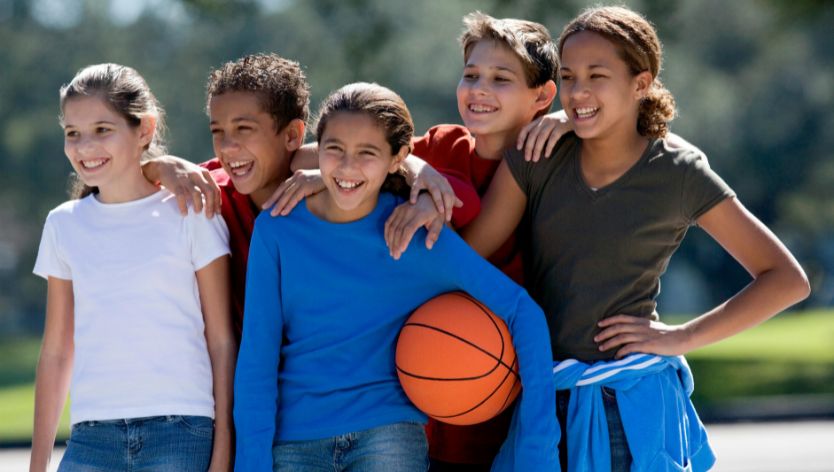 Five smiling children standing outdoors with their arms around each other, holding a basketball, representing teamwork and fun at a youth sports camp.