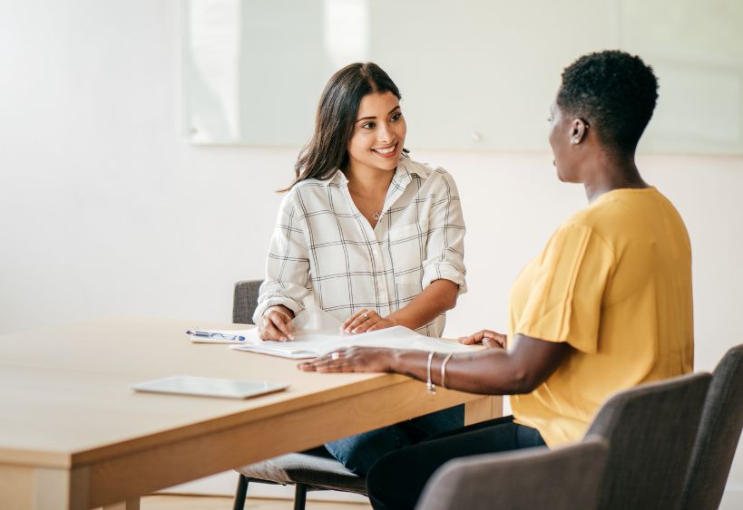 Two women sit at a table in a bright office, smiling and discussing documents during a professional meeting.