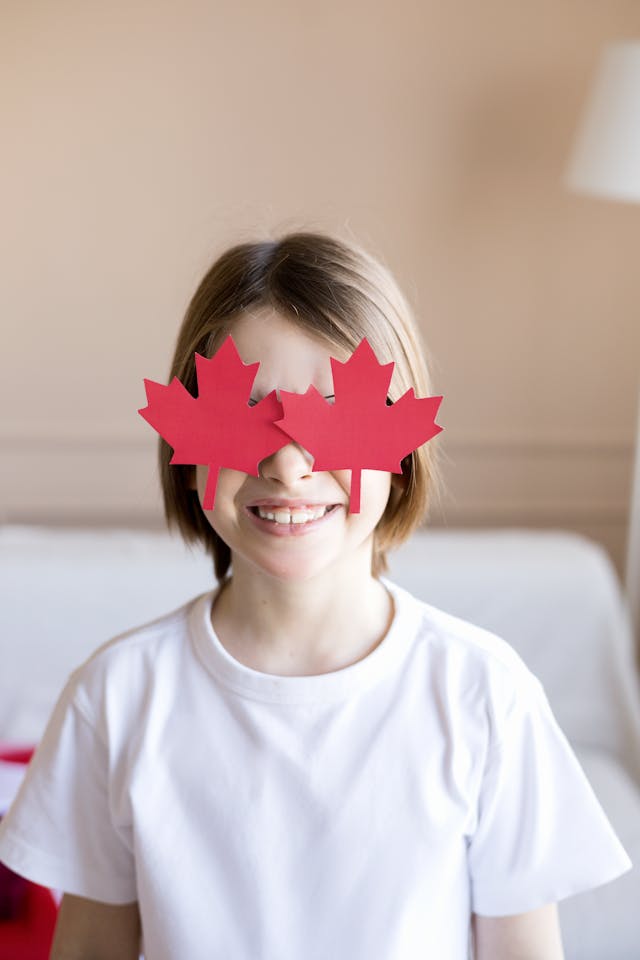 Smiling child with maple leaf glasses celebrating Canada Day indoors.