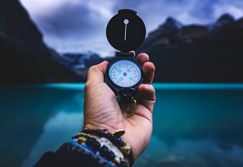 Outstretched hand holding a compass, guiding the way toward a tranquil lake framed by majestic mountains in the distance