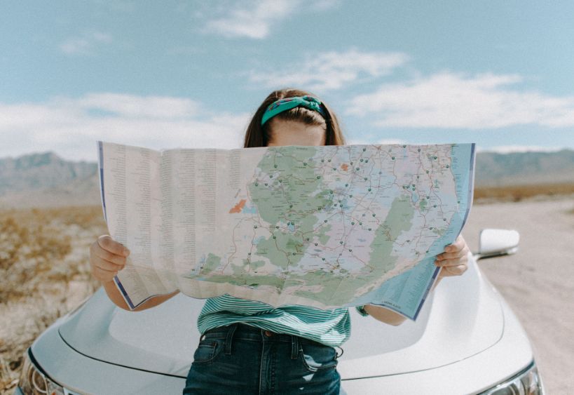 A female traveler with a map stands leans on the front of white car on a quiet road, mountains behind and possibilities unfolding under a clear blue sky