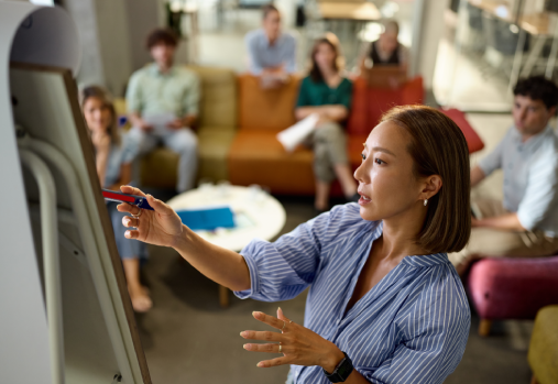 woman standing at the front of a room of people pointing to flip chart paper on an easel