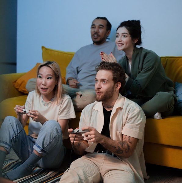 two people playing video games with two people sitting behind them on a couch watching 