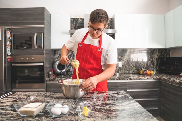 Teenager in a modern kitchen wearing a red apron while whisking ingredients in a stainless steel bowl.