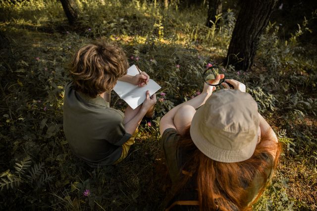 Two kids explore a forest, observing nature with a sketchbook and magnifying glass