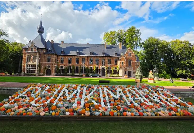 A historic manor house is pictured behind a large decorative display of colorful pumpkins arranged in a formal garden.