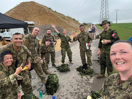 Group of CAF members in CADPAT smiling outdoors; backpacks and gear on ground. Overcast sky, dirt mound, and power lines in background.