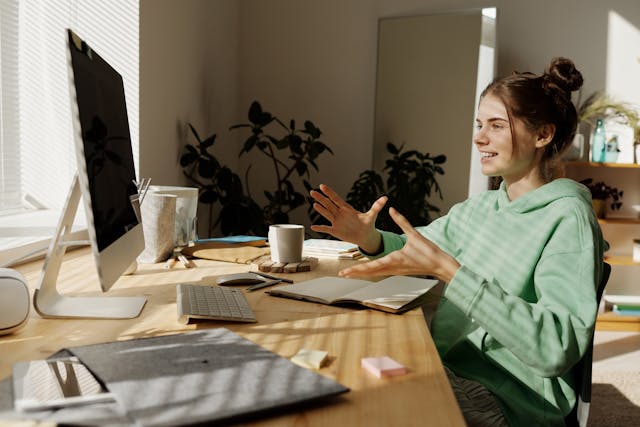 Young Woman in Green Hoodie Sitting Near the Table while Having Conversation Through Her Desktop