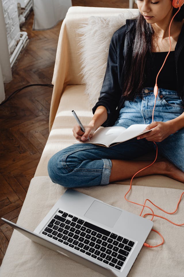 Student sitting on a couch writing in a notebook while a laptop is in front of her