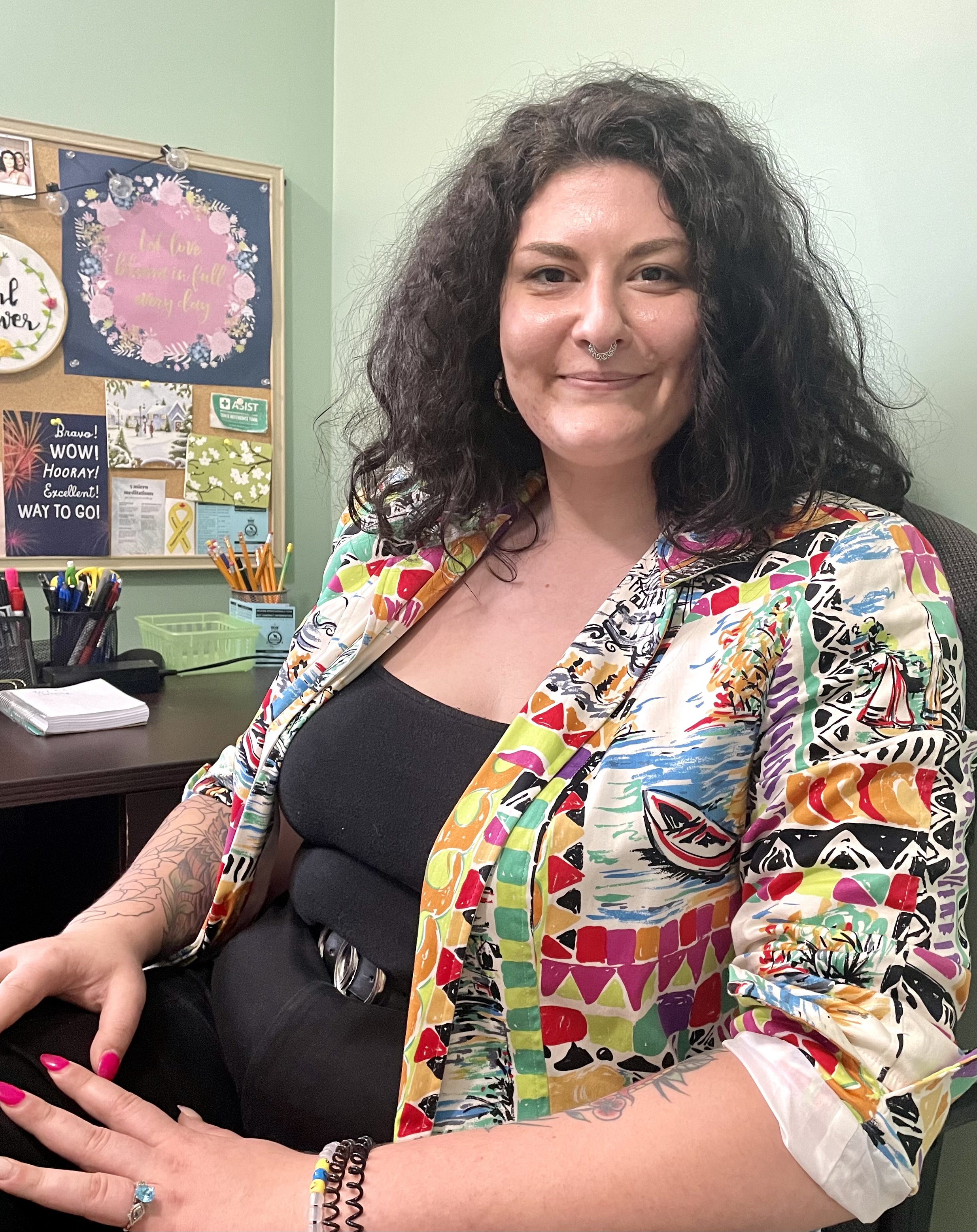 Young woman in black top and pants with a colourful coat, nose ring, tattoos, and dark pink fingernails