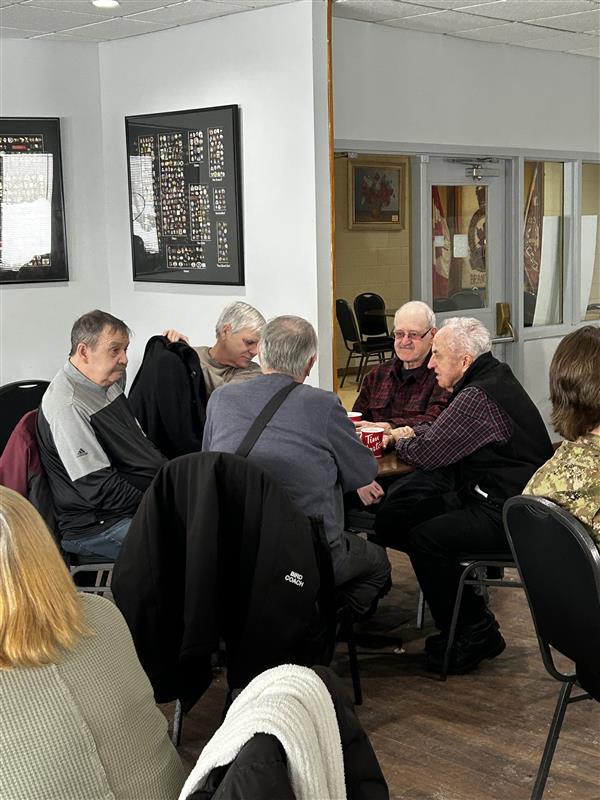 A small group of older Veterans seated around a table indoors, sharing coffee and conversation in a welcoming community space.