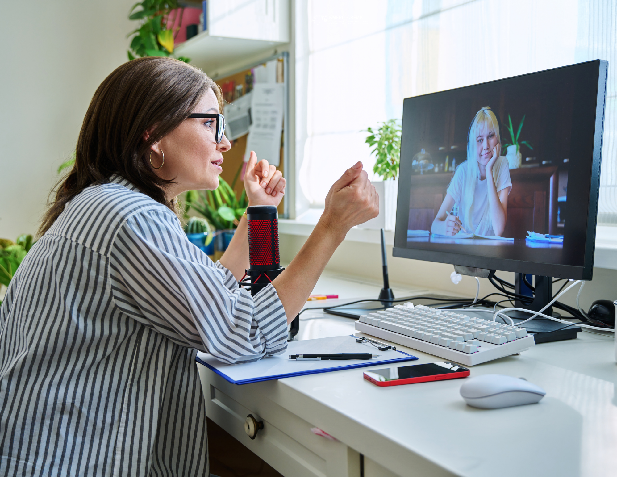 Person participating in a video call from a home office, speaking with someone on a computer screen in a bright workspace with plants and office equipment.