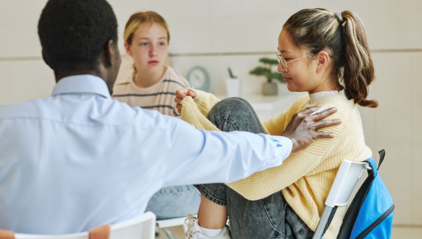Three youth sitting in a round and one reaching a supporting hand out to other 